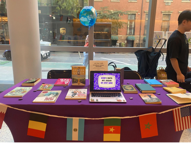 Table, covered in purple table cloth, various flags hanging in front (from right to left, U.S.A, Morroco,  Cameroon, Israel, and Belgium are visible). Table has multiple books on display as well as an open that has a message. There is a person behind the table to the right and behind the table is a window. 