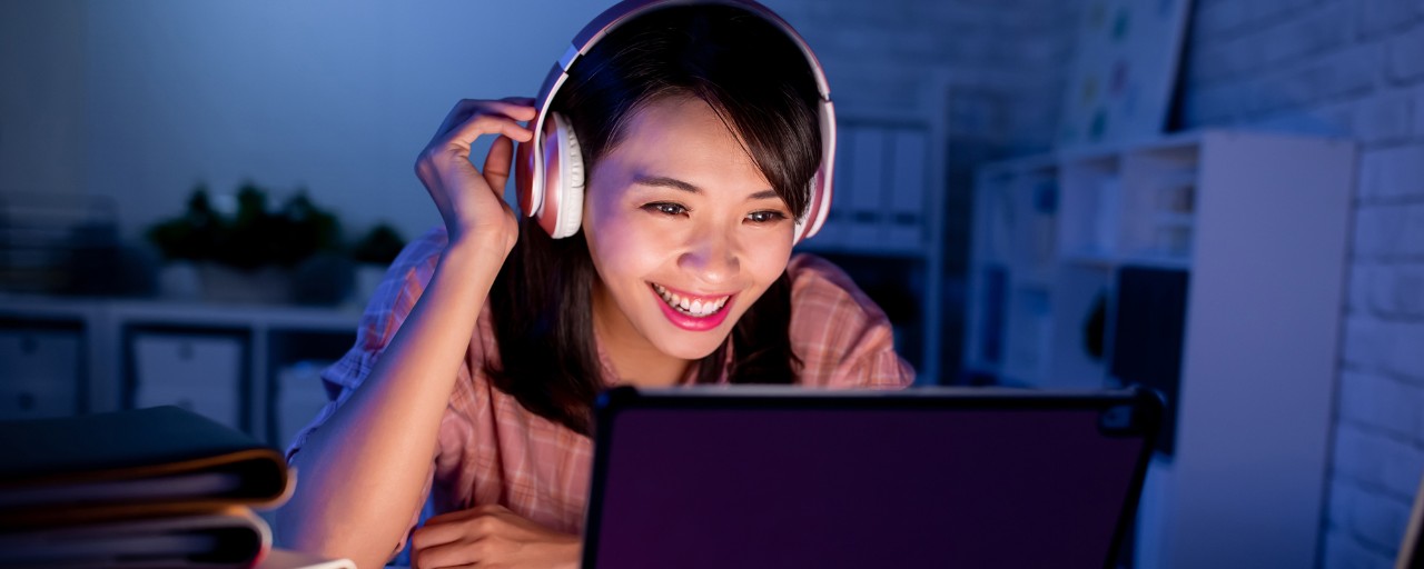 An online degree completion program student is wearing headphones and smiling while looking at a laptop screen.